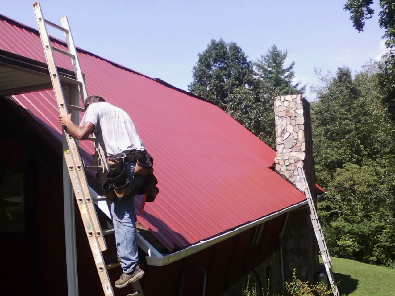 Roofer on a ladder beside a red metal roof and stone chimney — Soffit & Fascia Repair by Roofing Brothers in Ringwood